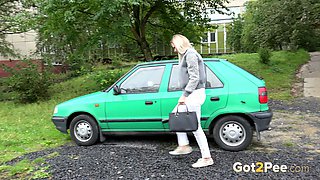 Bright blondie in white pants sits near the green car and pisses